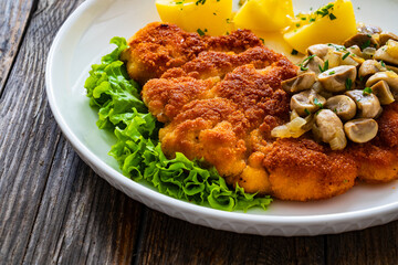 Crispy breaded seared chicken cutlet with fried white mushrooms and boiled potatoes on wooden table
