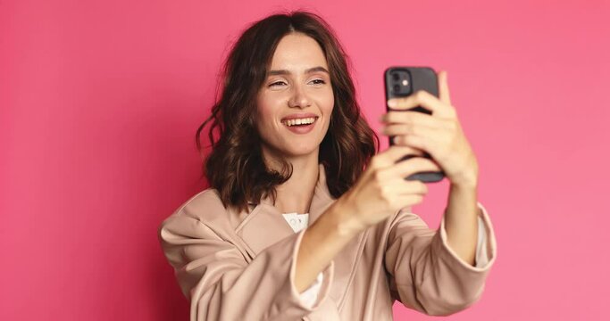 Happy girl stares at the phone and make photo. Female uses phone like mirror. Young girl make photo from hands with phone on pink background. Curly girl wear beige leather jacket, white hat and top.