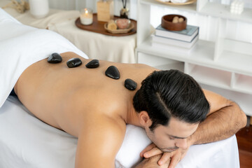 Fototapeta na wymiar Hot stone massage at spa salon in luxury resort with day light serenity ambient, blissful man customer enjoying spa basalt stone massage glide over body with soothing warmth. Quiescent