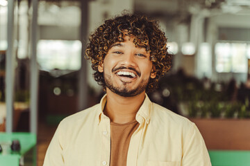 Portrait of smart smiling Indian student standing in university campus, education concept. Happy successful asian man wearing yellow casual shirt looking at camera in modern office 