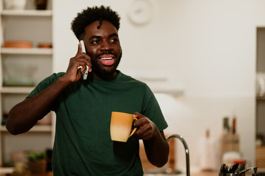 A cheerful interracial man is standing in a kitchen at home and talking on the phone