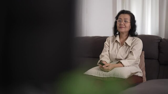 Senior woman holding remote control watching tv at home, recreation and entertainment.