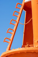 Fundament of sea buoy. A huge metal bright orange tube on the seashore. In the background, a light pavement, a wooden fence and the sea. Copy space. Background for quotes