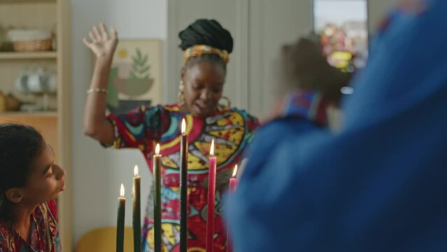Rack focus shot of African American man taking photos with phone of family on Kwanzaa celebration at home, cheerful wife posing and dancing while daughters looking at kinara candles