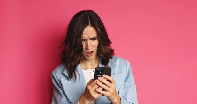 An angry brunette curly woman read some bad news or message and typing something on the phone and raising an eyebrow while standing in an pink studio.