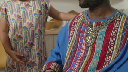 Tilt up shot of African American man wearing dashiki kaftan and kufi smiling and playing drums while little daughter dancing on Kwanzaa celebration at home