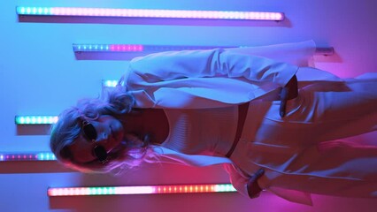 Vertical video. Young beautiful hipster woman in street style fashion concept. Hot model wearing white suit and sunglasses. Female posing in pink and blue neon light in studio interior - Powered by Adobe
