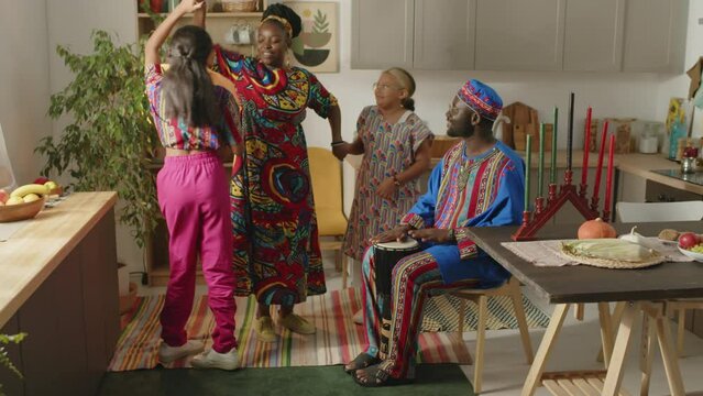 Full slow motion shot of African American father in dashiki clothing and kufi smiling and drumming while happy wife and two little daughters dancing on Kwanzaa home celebration