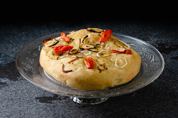 Focaccia with onion, cherry tomatoes, garlic and spices. Close-up, glass plate, on marble, granite table, black backgroun