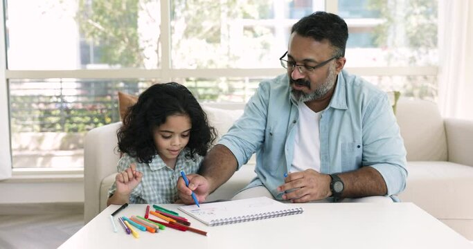 Senior 50s Indian man drawing pictures with felt-pens on paper sketchbook with little 5s grandkid, sit at table in living room, enjoy creative hobby spend leisure at home. Kids development, vocation