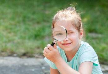 Child, young happy curious girl holding a magnifying glass next to her eye smiling, outdoors...