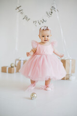 Fototapeta na wymiar Playful little girl wearing finest party dress standing alone in studio adorned with festive decorations. Charming caucasian baby girl with sincere emotions celebrating birthday party.