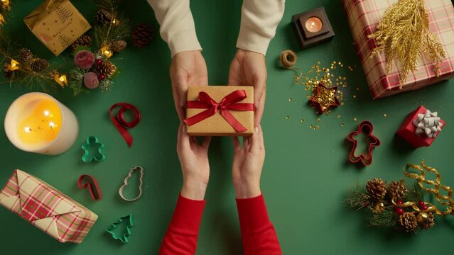 Giving a gift box for holiday to beloved people. Overhead view on lay flat green background with Christmas decorations, handsome male hands giving shiny golden glittery box with red bow to female 4K