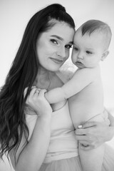 Black and white portrait of mom and nude beautiful daughter expressing pure and unconditional love between them. Charming woman standing at studio with little adorable toddler on hands.