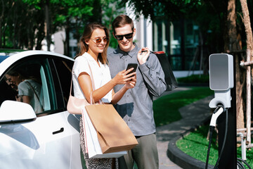 Young couple travel with EV electric car charging in green sustainable city outdoor garden in...