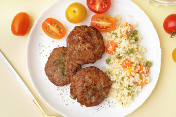Cutlets with bulgur, boiled vegetables and tomatoes on light yellow table in kitchen