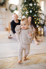 Focus on adorable baby girl in knitted wear running with barefoot at cozy apartment with festive decor. Joyful young parents sitting near Christmas tree on blurred background. Winter holidays concept.