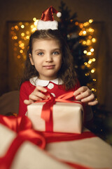 Fototapeta na wymiar 6 years old girl in Santa Claus costume wraps gifts boxes with red ribbons and festive lights on background
