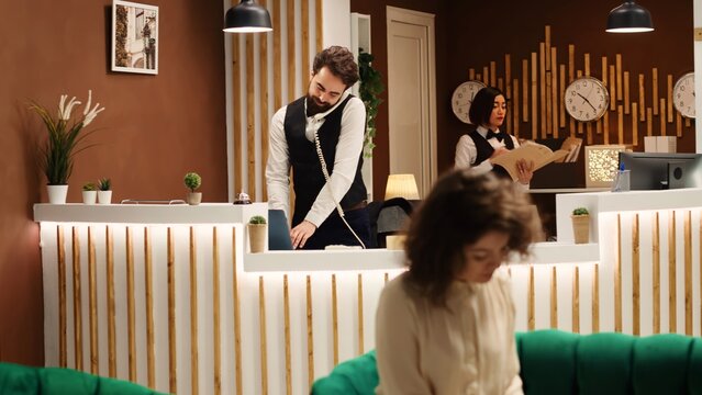 Helpful happy receptionist assisting tourists with picking hotel room and booking date via phone call. Teamworking concierge staff at their reception desk job in deluxe resort