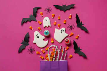 Shopping bag with tasty candy corns, cookies for Halloween and skeleton hand on pink background