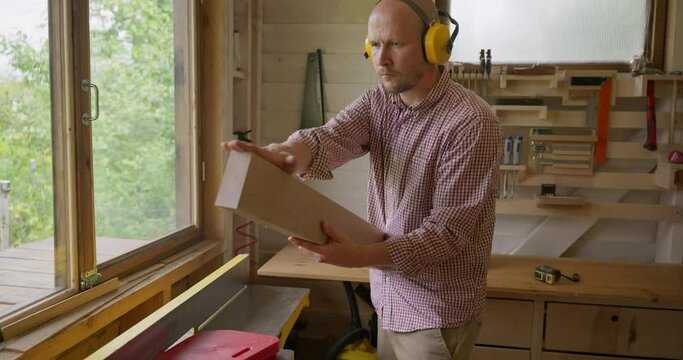 The middle plan is a side view. A male woodworker in protective headphones is whittling a board on a jointer machine in his workshop standing at a large bright window. High quality 4k footage