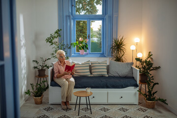 Obraz premium Elderly woman sitting on the sofa and reading a book