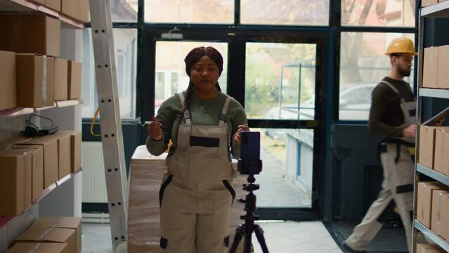 Warehouse chief supply chain officer using smartphone to make training video for interns. African american supervisor films herself in depository showing trainees how to seal cardboard boxes