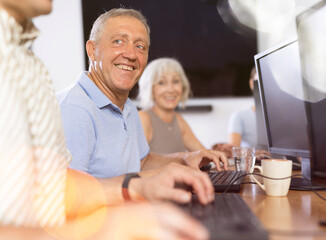 Fototapeta na wymiar Smiling motivated older man engrossed in exploring digital realm at internet cafe, using computer, demonstrating curiosity of modern seniors towards technology..