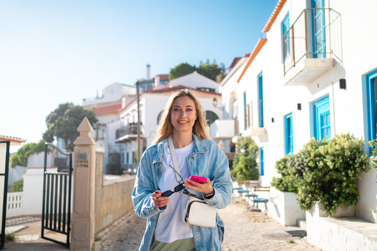 Woman walking down street in sunny coastal city. Attractive female with cheerful smile enjoys her vacation. Blond hair female tourist standing in street near cute tiny white houses. 