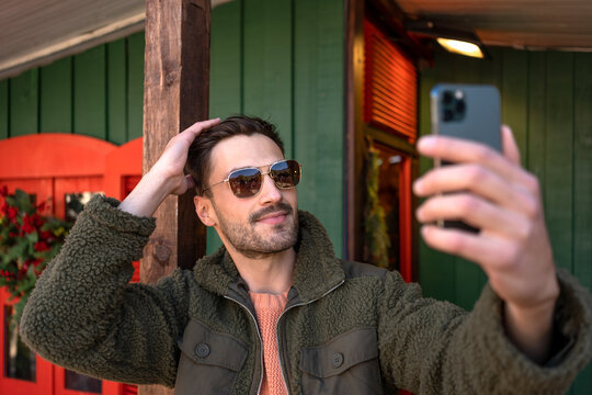 Man taking selfie outdoors. Stylish man wearing sunglasses making photo of himself with cell phone outside wooden house. Side view of handsome guy in fluffy jacket having video chat outdoor