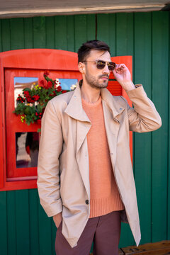 Handsome man outside Christmas house. Elegant brunette male touching his sunglasses. Man stands in front of green wooden house decorated for Christmas. Man in beige coat posing outdoors at winter