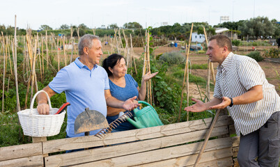 Neighbor conversation. Happy elderly couple chatting friendly with adult man while standing near wooden fence of country house vegetable garden in summer