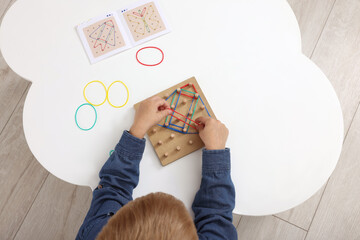 Motor skills development. Boy playing with geoboard and rubber bands at white table, top view