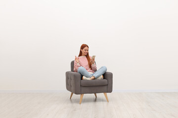 Happy young woman having video chat via smartphone while sitting in armchair near white wall indoors