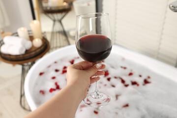 Woman holding glass of wine against bath tub with foam and rose petals indoors, closeup