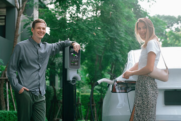 Young couple travel with EV electric car charging in green sustainable city outdoor garden in...