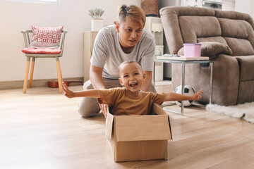 Happy Asian father driving cardboard box with excited little son inside. Father playing with kid at...