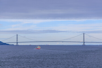 Small vessel sails calm blue waters by suspension bridge on cloudy day