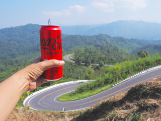Fototapeta premium Hand holding red can of Coca-Cola soft drink at viewpoint over curve street on mountains background.