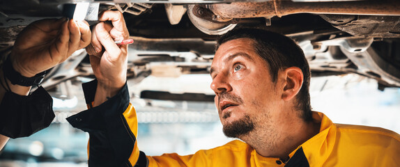 Vehicle mechanic conduct car inspection from beneath lifted vehicle. Automotive service technician in uniform carefully diagnosing and checking car's axles and undercarriage components. Panorama Oxus