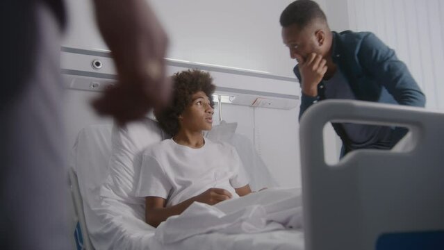 Man visits son in hospital ward and discusses results of operation. Nurse talks with African American boy and raises back of his bed. Patient recovering after successful surgery. Modern bright clinic.