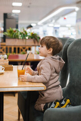 Four year old boy sits at a table in a cafe and looks at a glass of orange juice, holding a straw in his hands