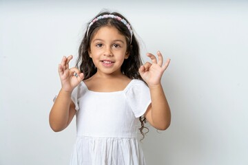 beautiful kid girl wearing white dress showing both hands with fingers in OK sign. Approval or...