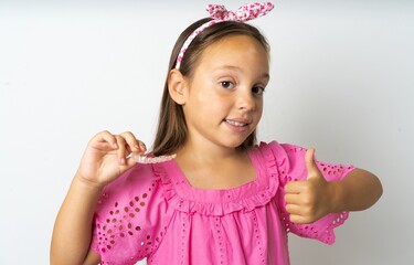 beautiful kid girl wearing pink dress holding an invisible braces aligner and rising thumb up, recommending this new treatment. Dental healthcare concept.