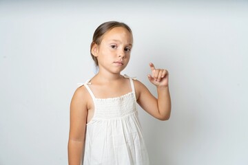 Displeased beautiful kid girl wearing white dress shapes little hand sign demonstrates something...