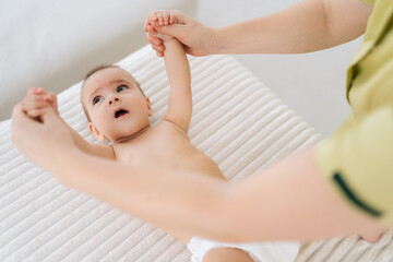 Top view of happy infant baby boy lying in bed playing with mom at home. Caring young mother doing gymnastic exercises with kids arms. Concept of babysitting, maternity leave motherhood babyhood.