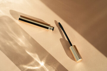 Mascara brush lies next to an open tube, on a beige background with shadows from sunlight. Copy...