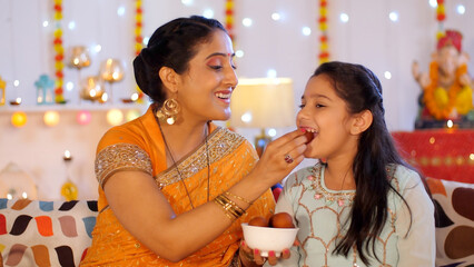 Indian mother and daughter celebrating Indian Hindu festival Diwali at home - colorful background. A cheerful middle-aged lady is feeding her young daughter sweets during festivities - celebration ...