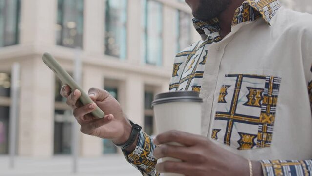 Handsome young Black man wearing fancy patterned shirt standing with takeaway coffee cup outdoors in street and browsing on smartphone