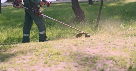 Worker with manual petrol lawn mower mows green grass in city park. Landscaping, work on ennoblement of public place